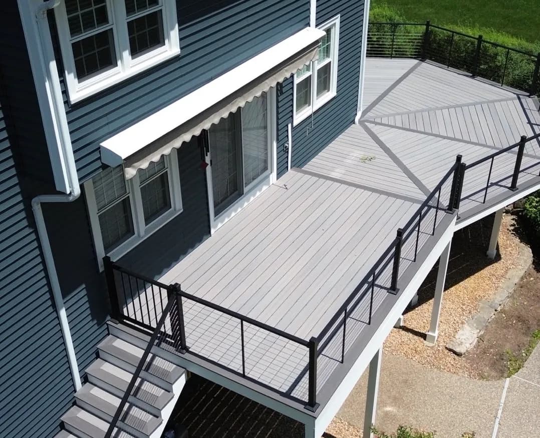Elevated gray composite deck with a picture-frame border and diagonal inlay, black horizontal-cable railing, and stairs down to grade