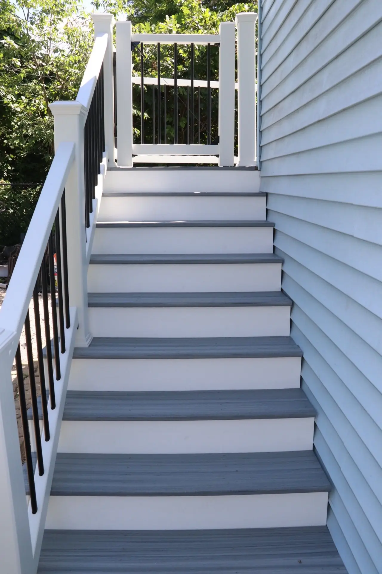Stair run with gray composite treads and white risers climbing beside light blue siding, with a white railing, black balusters and a gate at the top