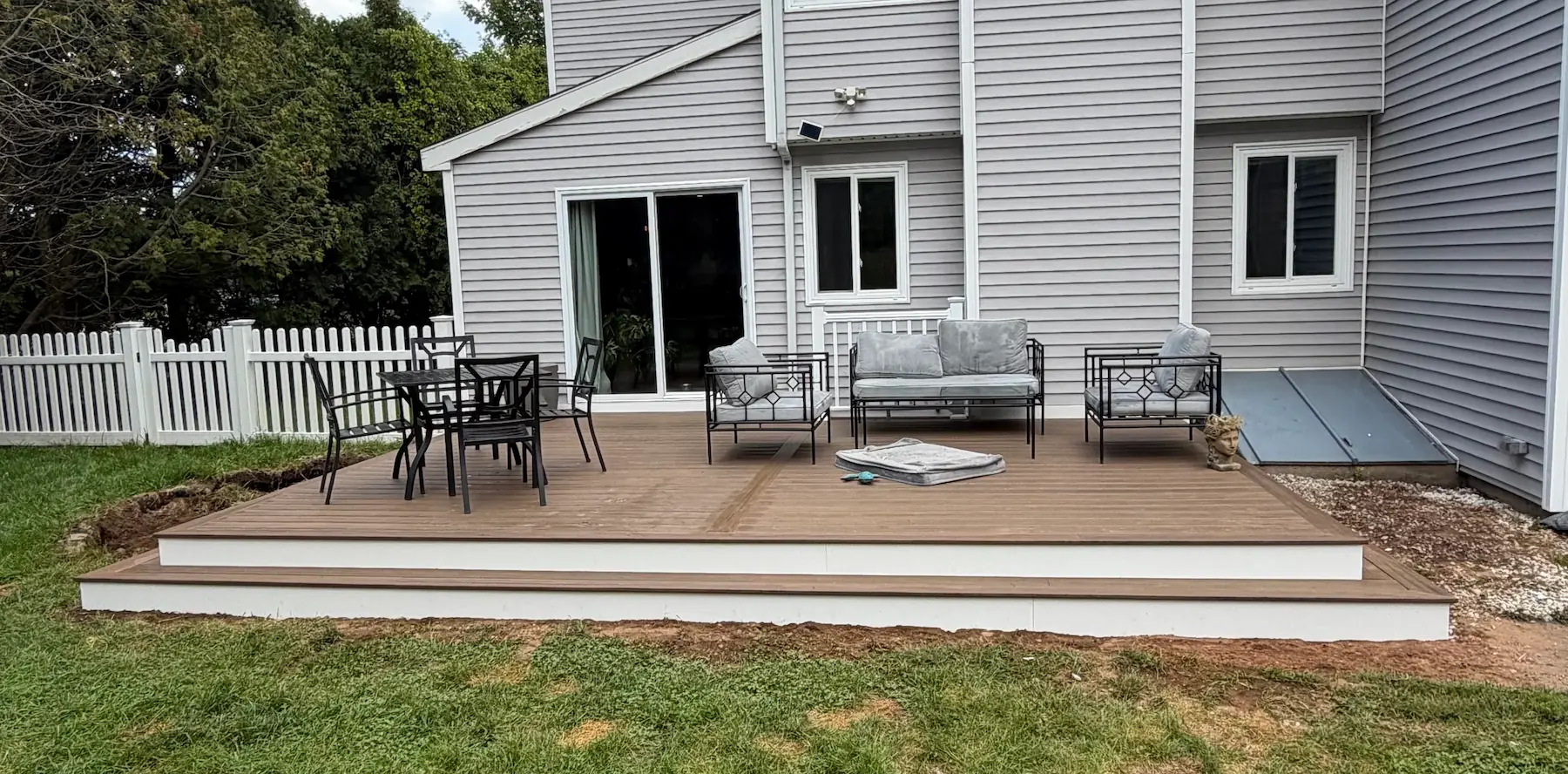Ground-level composite platform with a stepped border and white riser fascia, set with outdoor furniture against a gray clapboard house