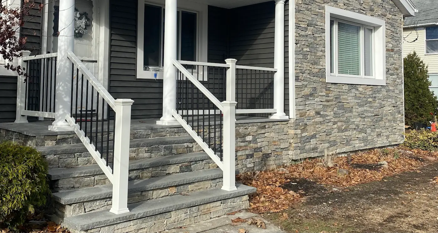 Covered front porch with white round columns and white PVC railings with black balusters, over stacked-stone steps with bluestone treads beside a stone veneer wall