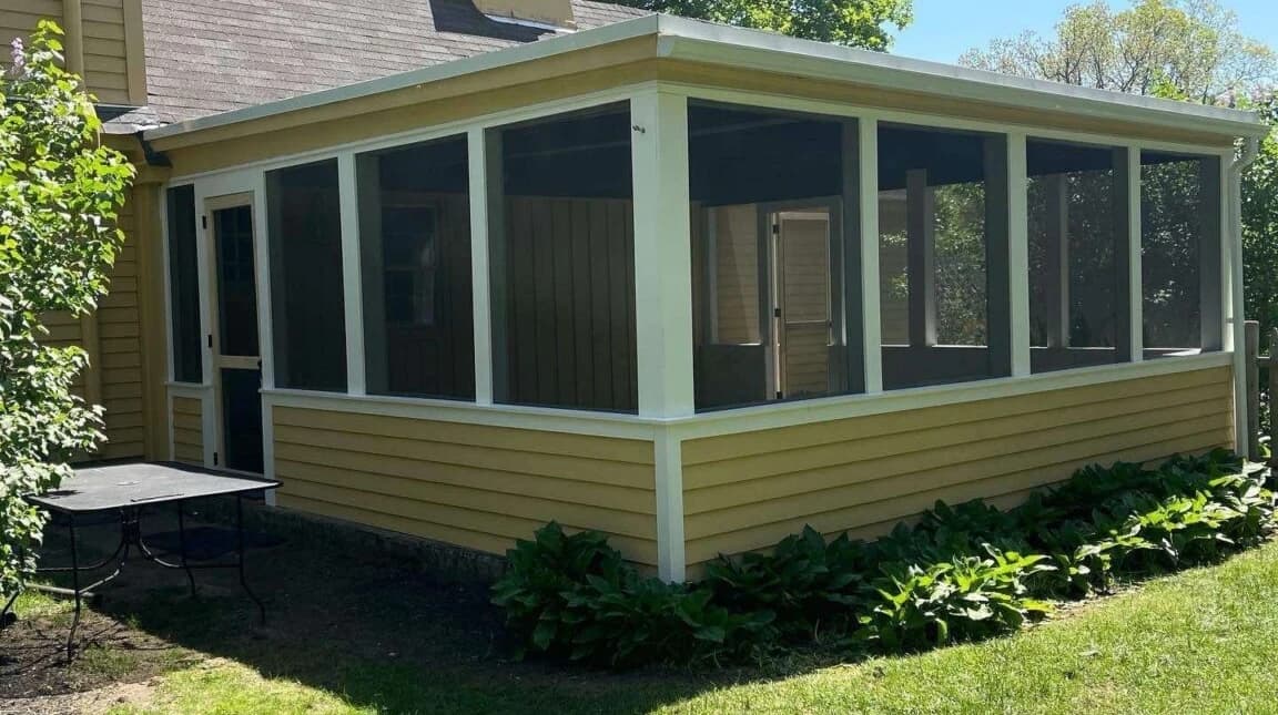 Ground-level screened-in porch with white trim and full screen panels, built onto a house with yellow clapboard siding