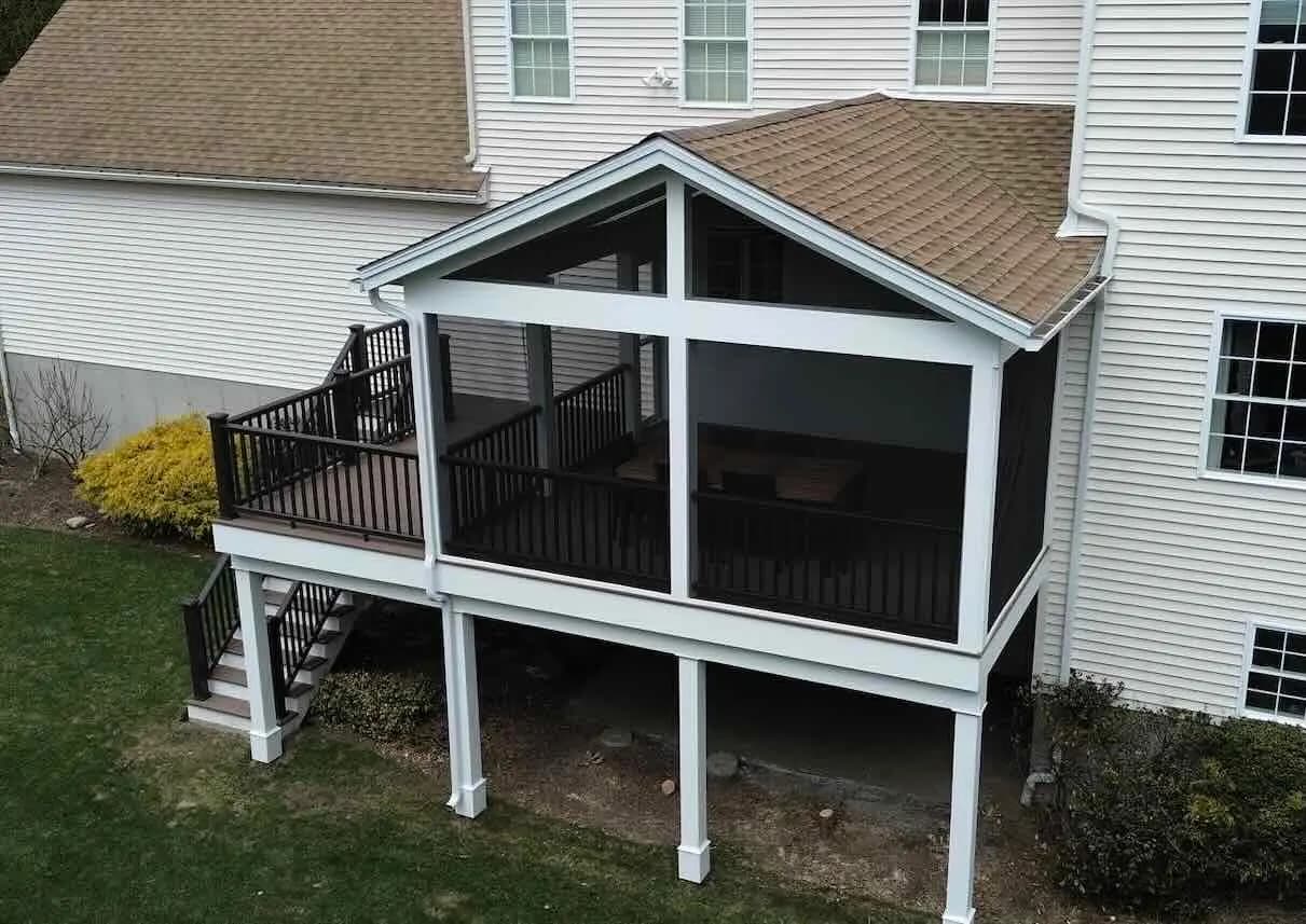 Elevated screened-in porch under a gable roof, with white PVC posts and dark railings, joined to an open deck with stairs down to the lawn